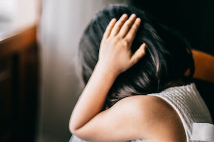 Young girl in emotional distress, sitting indoors with hand on head.